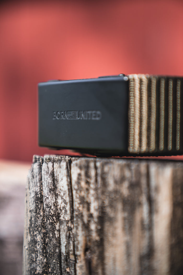 A close-up of the BU Logo Belt - Army Green by Born United Private Label, featuring a men's ratchet buckle engraved with “BORN UNITED,” resting on a weathered wooden post against a blurred red background.