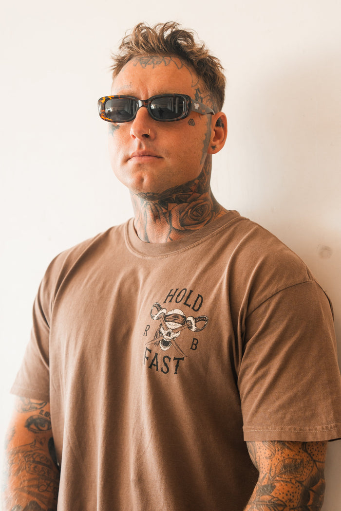 A man with neck and arm tattoos wears dark sunglasses and a Relentless Betrayal Hold Fast Premium Cocoa T-Shirt featuring an anchor graphic. Made from heavyweight jersey cotton, he stands confidently against a plain, light background.