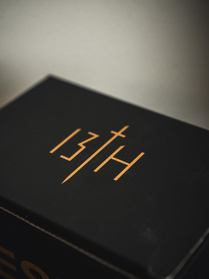 A black 13th box, featuring gold "13TH" with a cross-shaped "T," sits next to a Jesus Saves Mug by 13th. Both are set against a blurred neutral background.
