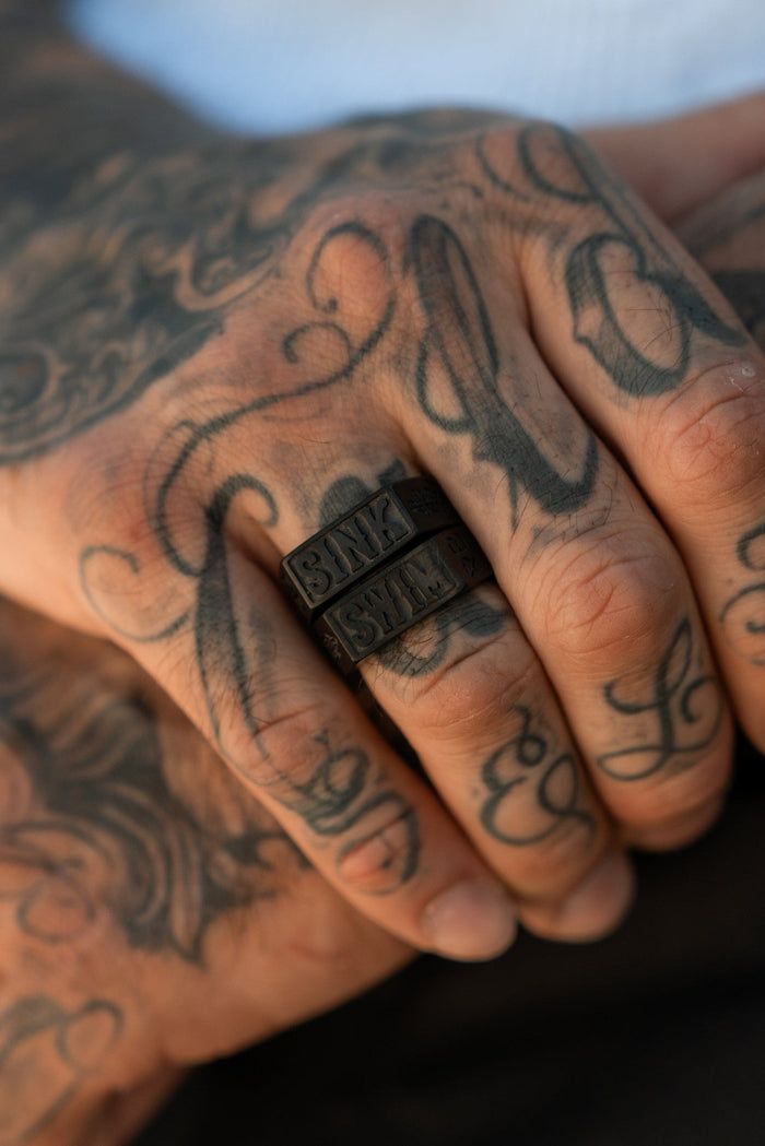 A tattooed hand showcases the Relentless Betrayal Matte Black Sink or Swim Rings, featuring SAVE THEM inscriptions on two bands. The stainless steel rings add edgy elegance against a blurred background, with tattoos of swirling patterns and letters enhancing the look.