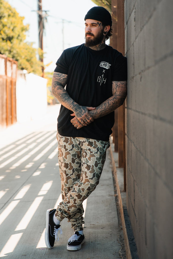 A tattooed man in a black beanie and tee stands outdoors, wearing Mucker Bucket Men's Classic Premium Joggers in Duck Camo and black sneakers, hands clasped, ready for running or relaxing.
