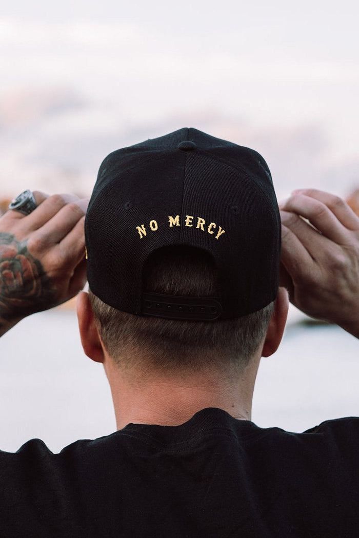 A person wearing a Relentless Betrayal No Quarter snapback adjusts the cap with both hands. No Mercy is written in gold on the back. A tattoo adorns their hand, and a ring glints on one finger, while a blurred background highlights their style.