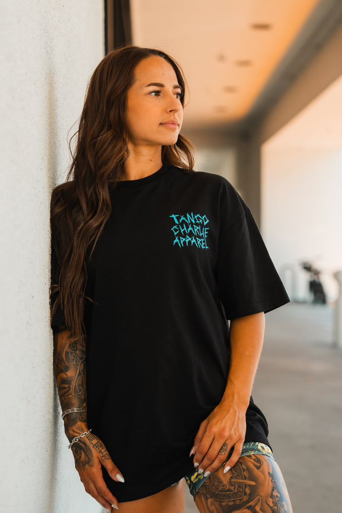 A woman with long brown hair and arm tattoos leans against a white wall in the sun, wearing the Tango Charlie Apparel Psych Ward Premium Oversized Tee—black cotton with Tango Charlie Apparel in blue lettering. She gazes to the side.