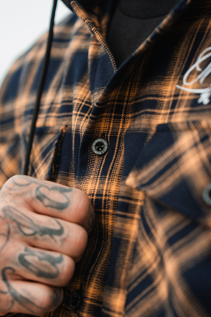 A close-up features tattooed hands gripping the collar of a Relentless Betrayal Skilled Sailor Premium Hooded Flannel, highlighting its ultra-soft cotton/polyester blend. The brown plaid shirt, with black buttons and an embroidered chest pocket, completes the look.