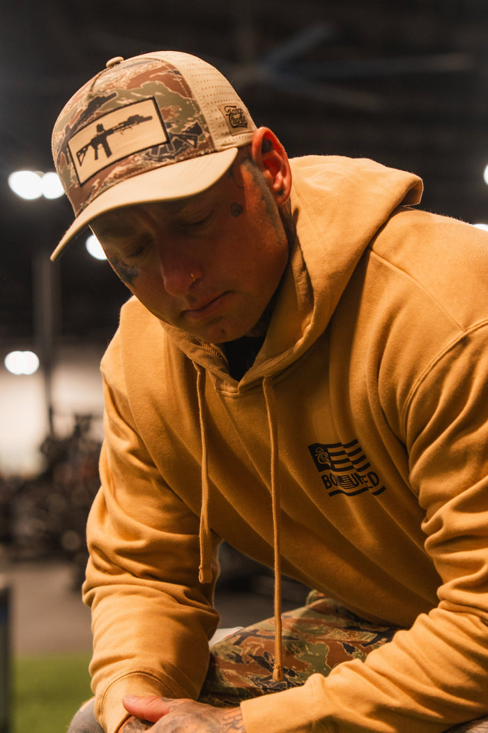 A person in a tan hoodie, camouflage pants, and a Tango Charlie Apparel Snipes & Stripes Hydro - Snapback Hat featuring a rifle graphic gazes downward indoors, with blurred gym equipment in the background.