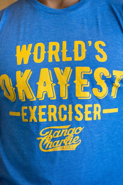 A person wears a comfy blue cotton/poly blend tee with yellow WORLDS OKAYEST EXERCISER text from Tango Charlie Apparel, set against a blurred wooden background with tattoos on their arm.