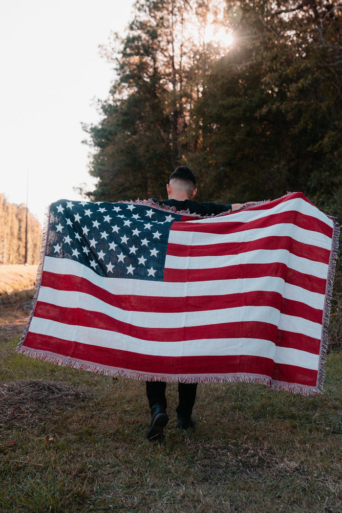 A person walks away on a grassy path, holding an American Flag Jacquard Blanket from Born United Private Label. The sun peeks through tall trees in the background, casting a warm glow on this quintessentially American scene.