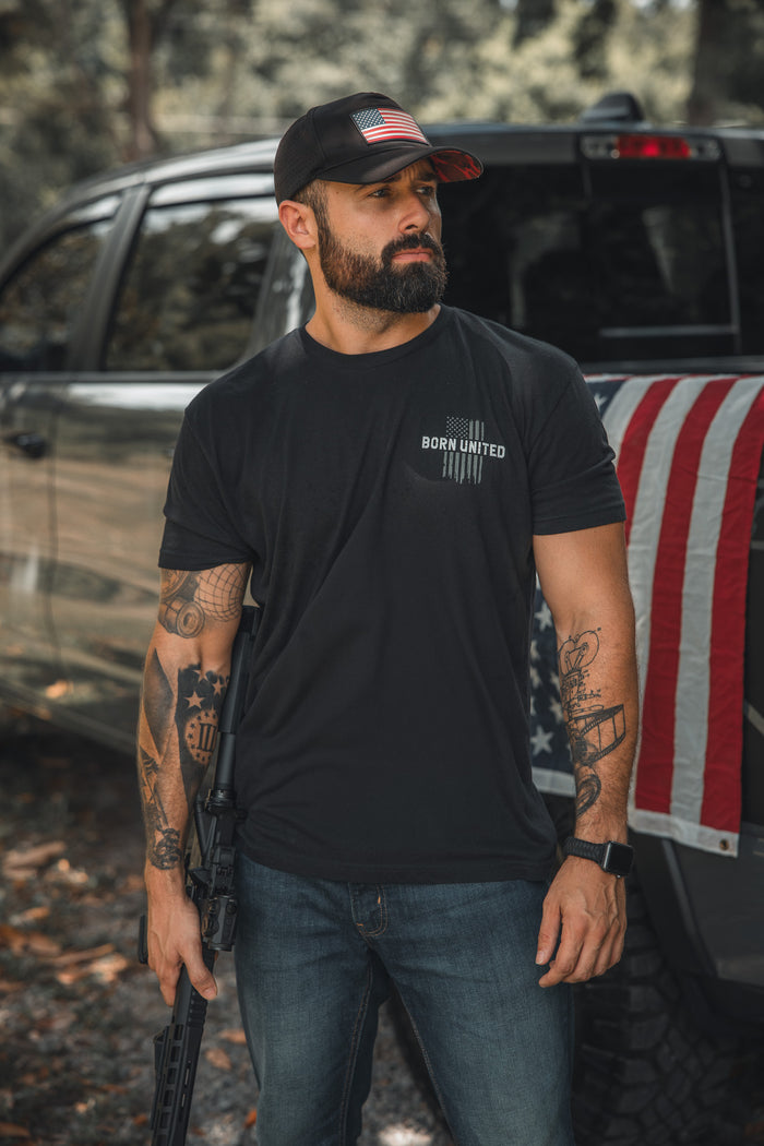 A tattooed man wearing the Born United Private Label American Heroes Tee and an American flag cap stands by a truck with an American Heroes Flag over the tailgate, holding a rifle and looking serious outdoors.
