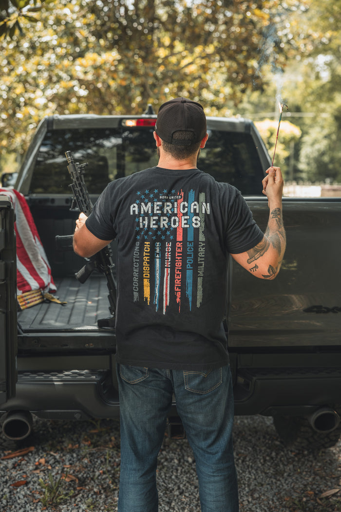 A man stands in a pickup truck holding a rifle and sparkler, wearing the Born United Private Label American Heroes Tee. The shirt features stripes and words like Honor and Sacrifice. An American flag is draped nearby.