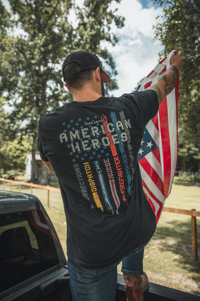 A man stands in a pickup bed holding an American Heroes Flag, wearing the American Heroes Tee by Born United Private Label. The black shirt features colorful stripes honoring corrections, dispatch, fire, EMS, law enforcement, and military.