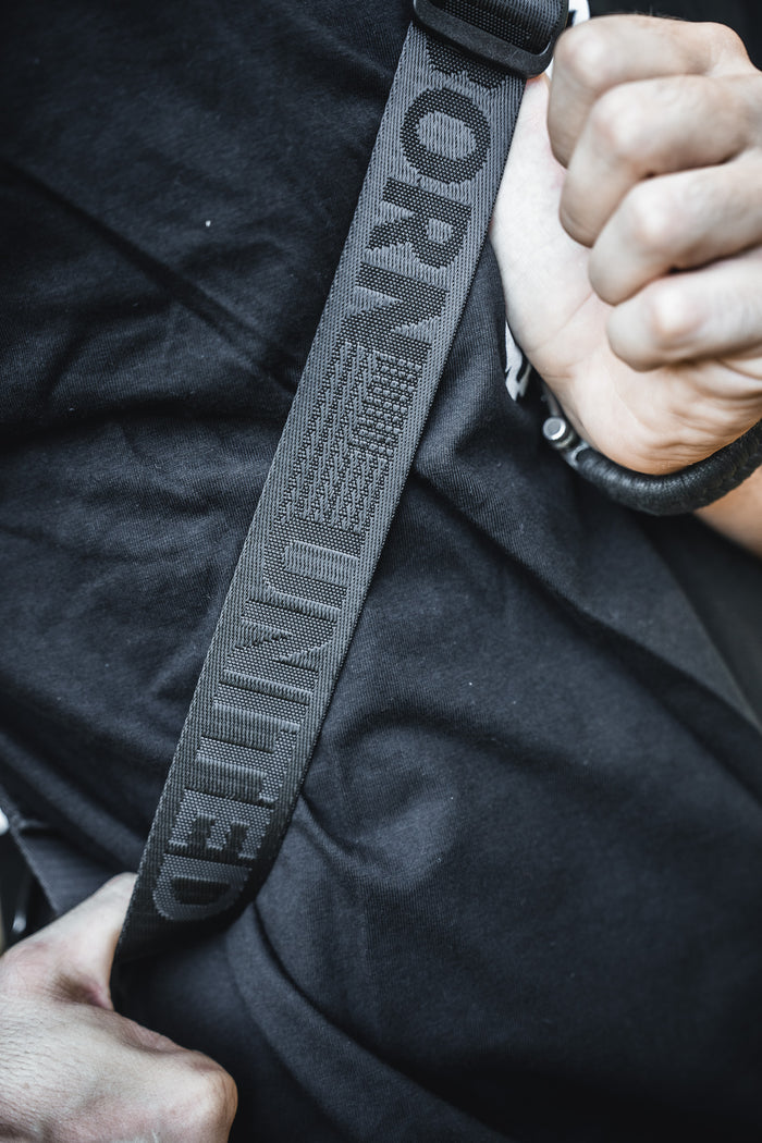 A close-up shows a person in a black shirt fastening a seatbelt marked "UNITED" and sporting the stylized flag. They carry the Born United Private Label Black Phantom Tactical Sling-Pac - 4L for easy concealed carry and daily essentials.