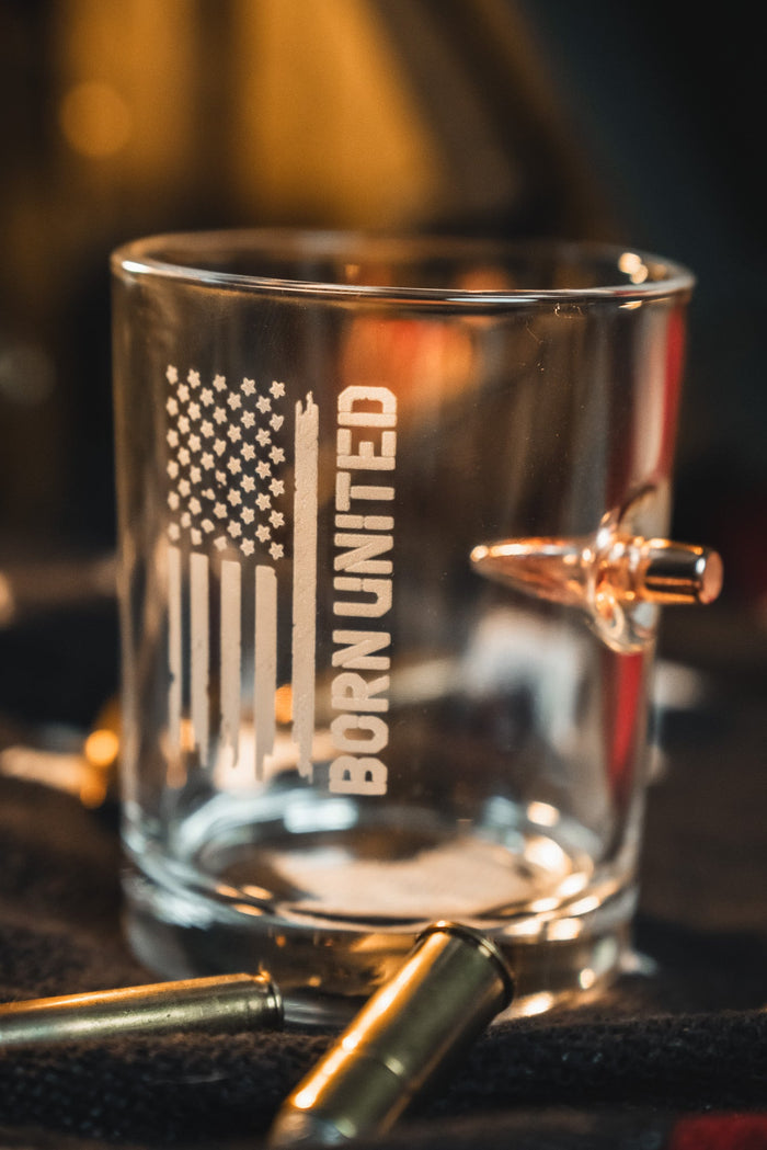 The Born United Bullet Whiskey Glass by Born United Private Label, featuring a U.S. flag, “BORN UNITED,” and an embedded bullet, rests amid scattered bullets on a dark surface.