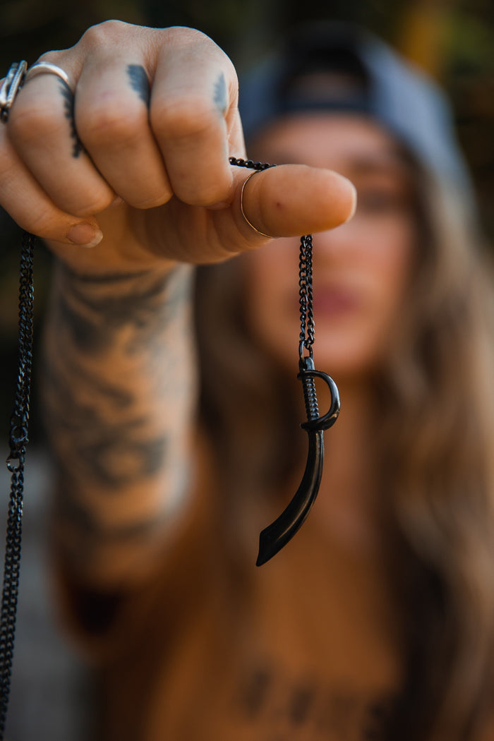 A tattooed individual wearing a cap presents the Relentless Betrayals Broadsword Necklace, featuring a handcrafted black pendant shaped like a curved blade on a stainless-steel chain. Ring-adorned fingers add intrigue as the blurred background emphasizes this striking piece.