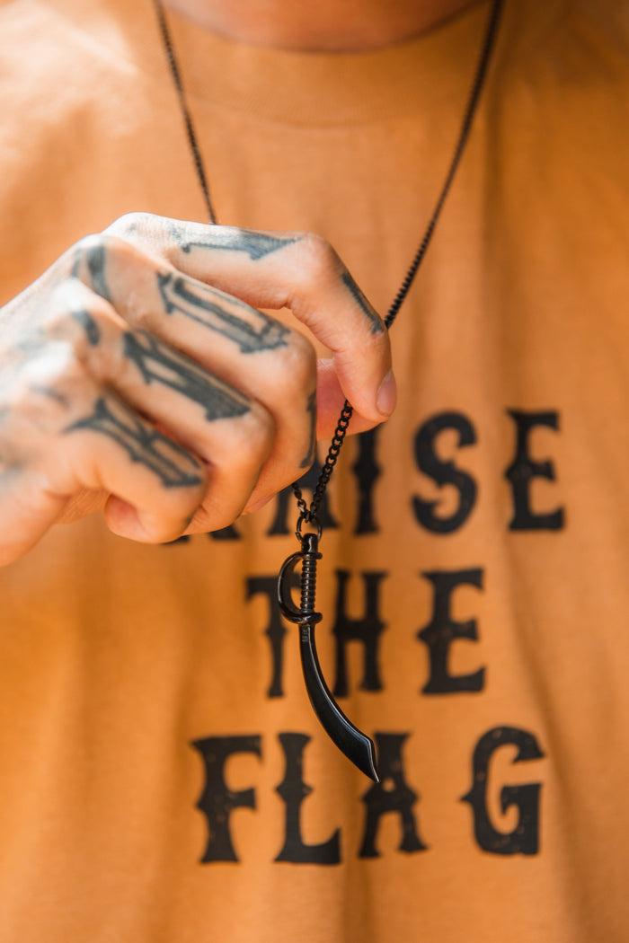 A tattooed hand holds a Broadsword Necklace by Relentless Betrayal, featuring a broadsword pendant on a stainless-steel chain. The person wears a mustard shirt with partially visible RAISE THE FLAG text.