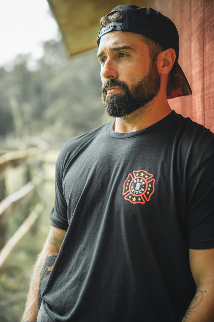 A bearded man in a backward black cap and Born United Private Labels Brotherhood Forged In Flames T-shirt stands outdoors by a wooden fence and red building, gazing thoughtfully into the distance.