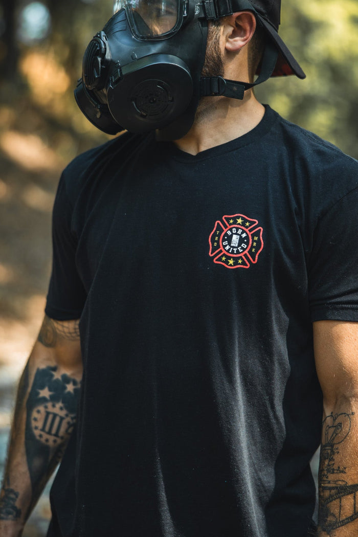 A tattooed individual stands outdoors in a Born United Private Label Brotherhood Forged In Flames black T-shirt, black cap worn backwards, and a gas mask—symbolizing firefighting safety.