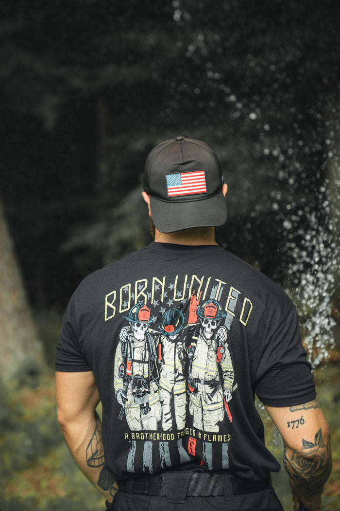 A person outdoors wears a Born United Private Label Brotherhood Forged In Flames black T-shirt with three firefighters and a black cap featuring a U.S. flag patch.