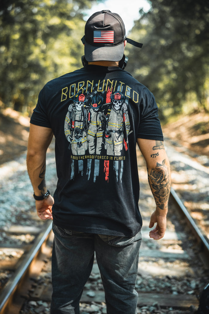 A man stands on railroad tracks, wearing a Born United Private Label black cap with a U.S. flag patch and the Brotherhood Forged In Flames shirt, featuring three firefighters, BORN UNITED, and an American flag. Trees line the tracks behind him.