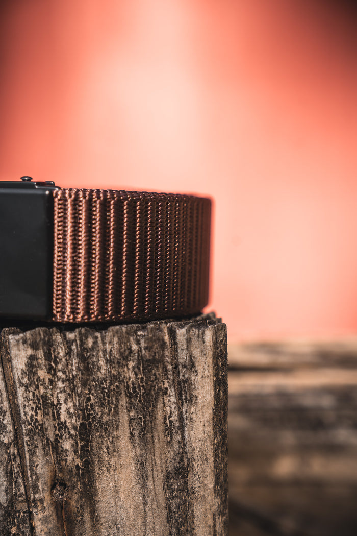 Close-up of the BU Logo Belt - Brown by Born United Private Label, featuring an adjustable design, resting on a weathered wooden surface with a blurred peach-colored background.