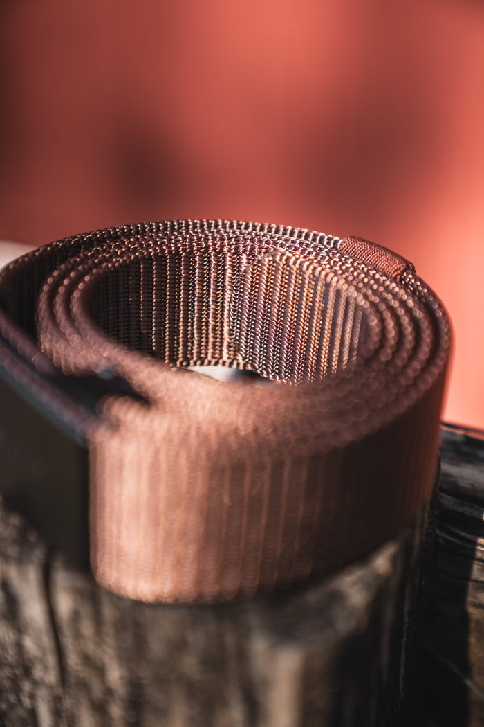 A close-up of the BU Logo Belt - Brown by Born United Private Label, an adjustable men's brown nylon belt, rolled up on a wooden surface with a blurred reddish background.