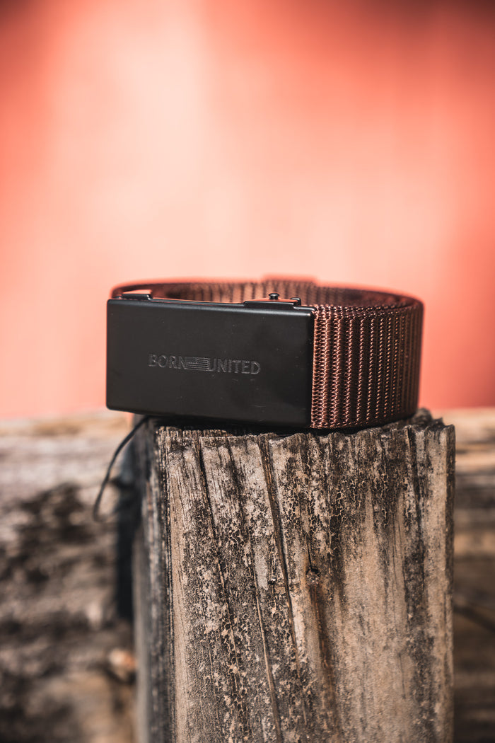 The BU Logo Belt - Brown from Born United Private Label is a men's brown woven fabric belt with a black buckle and adjustable fit, coiled on a weathered wooden post against a blurred reddish background for style and comfort.