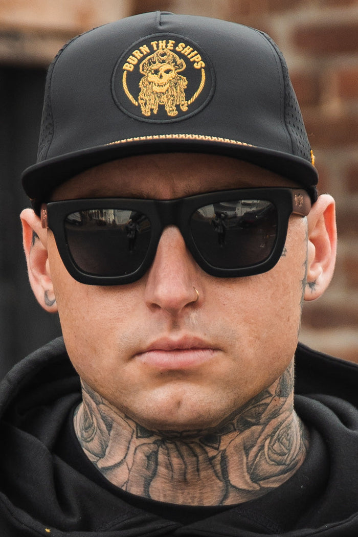 Sporting tattoos and facial piercings, wearing the Relentless Betrayals Burn The Ships BLACKOUT Snapback featuring a detailed gold emblem, black sunglasses, and a black hoodie. A brick wall serves as the backdrop.