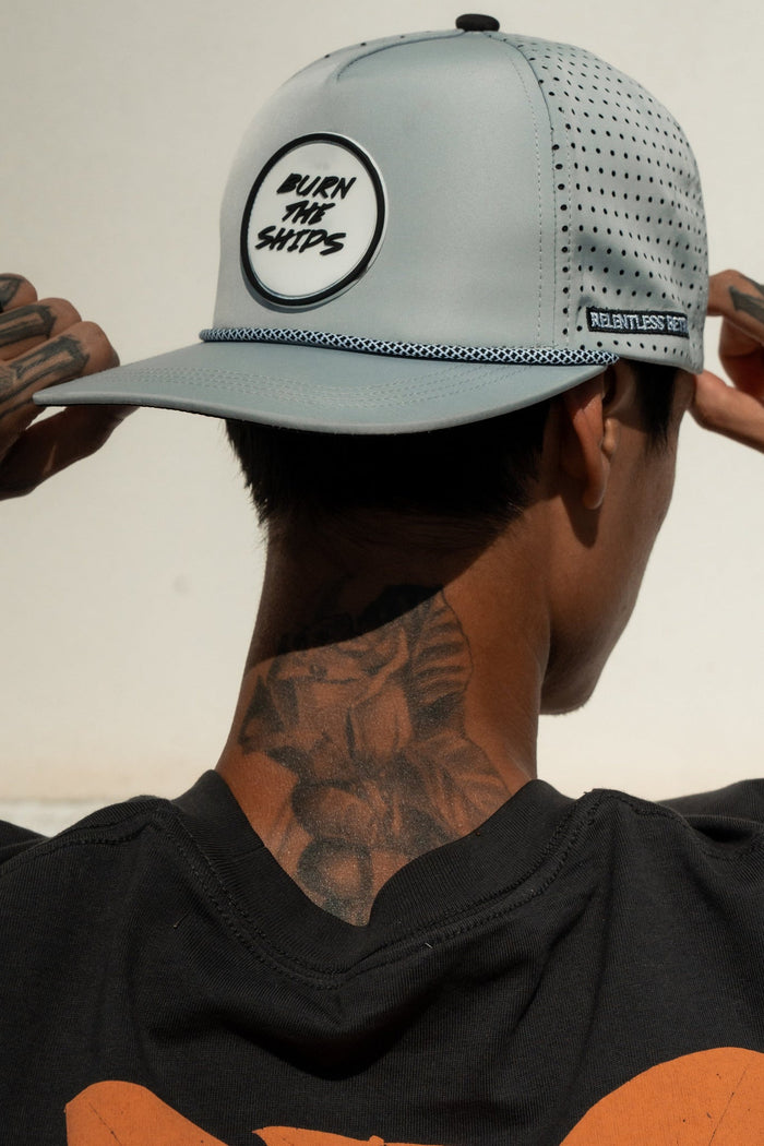 A person adjusts the Relentless Betrayal Burn The Ships Cement PVC Snapback, fitting the pirate lifestyle with its PVC rubber patch accents and ventilation holes. Paired with a black shirt, tattoos on neck and hands enhance their rugged allure.