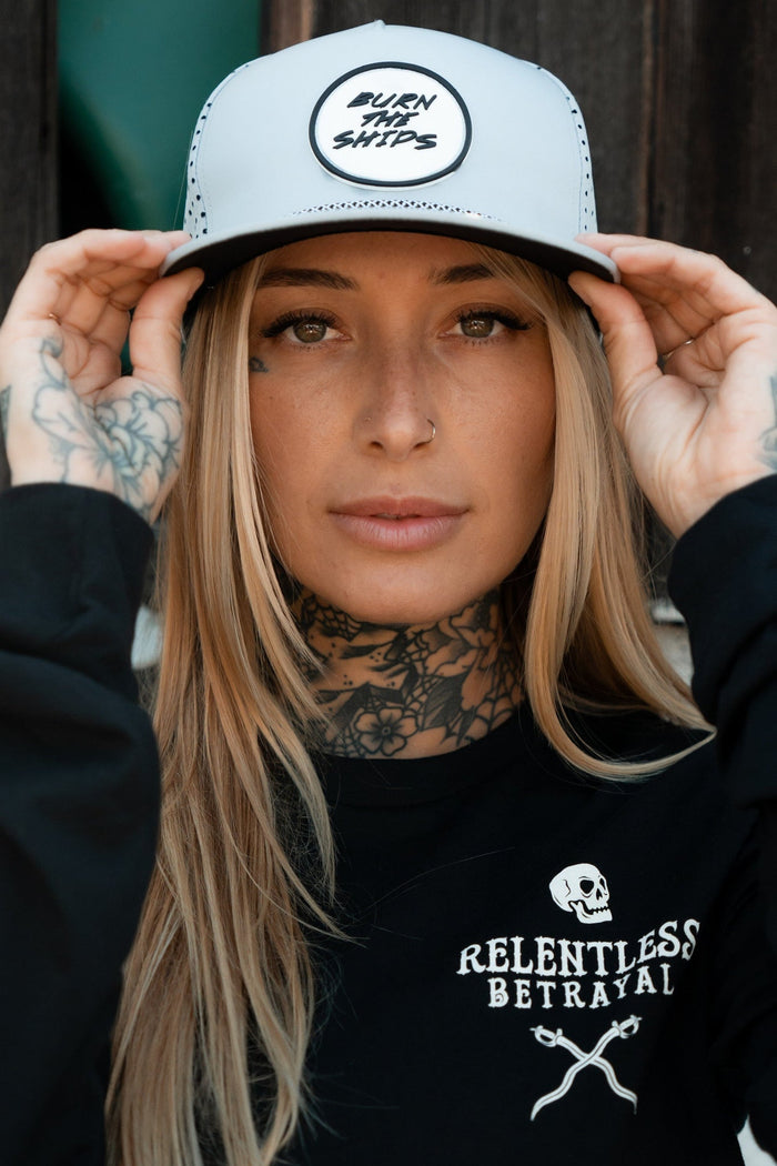 A person with long blonde hair and tattoos adjusts the brim of a Burn The Ships Cement PVC Snapback by Relentless Betrayal. Theyre wearing a black shirt featuring white graphics, including a skull, and the text Relentless Betrayal, embracing a pirate lifestyle.