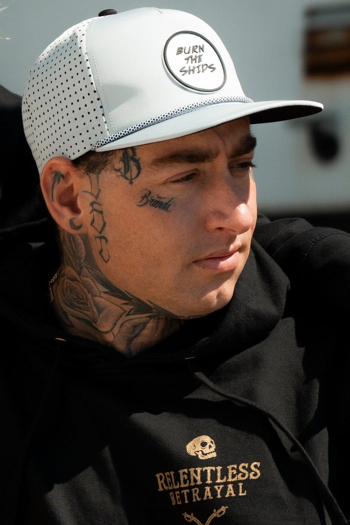 Wearing the Burn The Ships Cement PVC Snapback and a Relentless Betrayal black hoodie with a skull design, tattoos on their face and neck, they embody the pirate lifestyle outdoors, gazing into the distance.