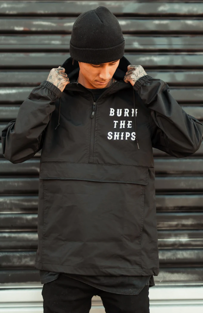 In front of a corrugated metal backdrop, a person adjusts the hood of their unisex Burn the Ships Pullover Rain Jacket by Relentless Betrayal. The black jacket, emblazoned with BURN THE SHIPS, boasts a water-resistant coating and complements their tattooed hands.