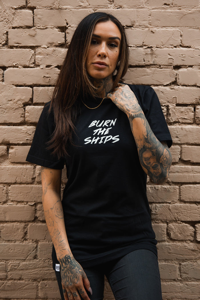Against a brick wall, a long-haired person with tattoos wears the Burn The Ships Traditional Tee by Relentless Betrayal. This black unisex tee, crafted from fine jersey cotton with durable double stitching, features the text BURN THE SHIPS. One hand rests on their neck and the other hangs down.