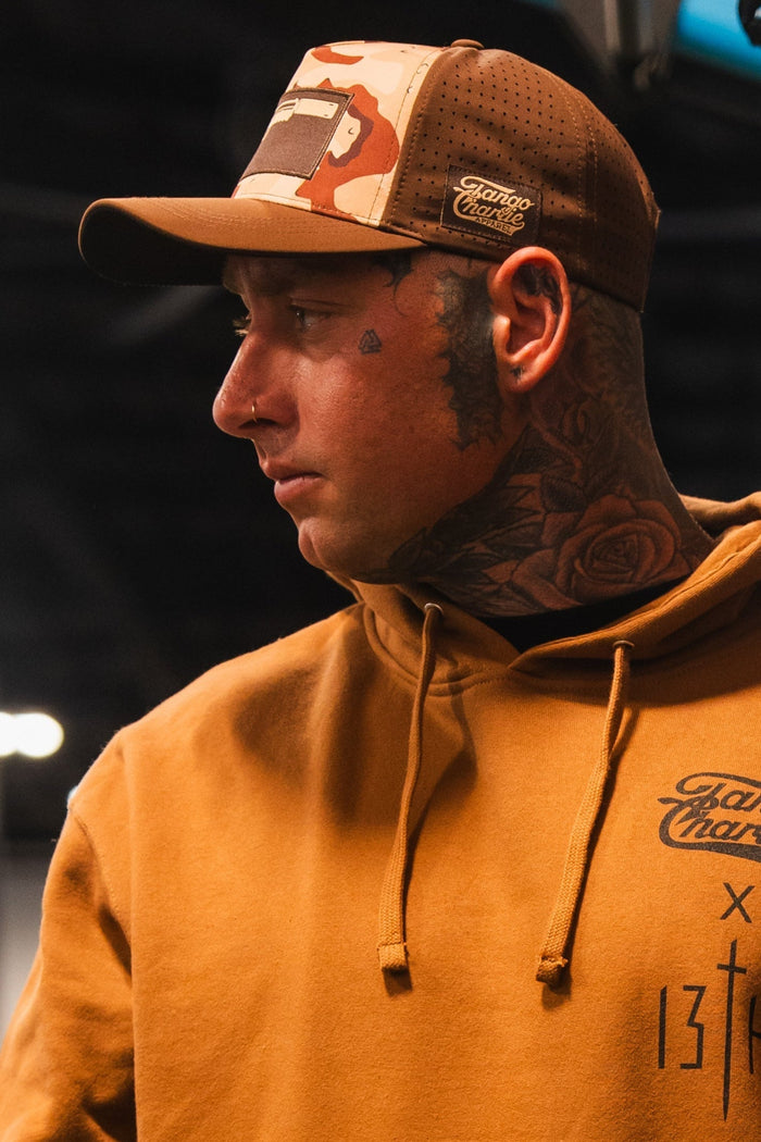 A man in a brown hoodie and a Tango Charlie Apparel Choco Chip Silencer Hydro Snapback Hat looks to the side, revealing neck and face tattoos—a rose and teardrop—and a nose ring, with a blurred background.