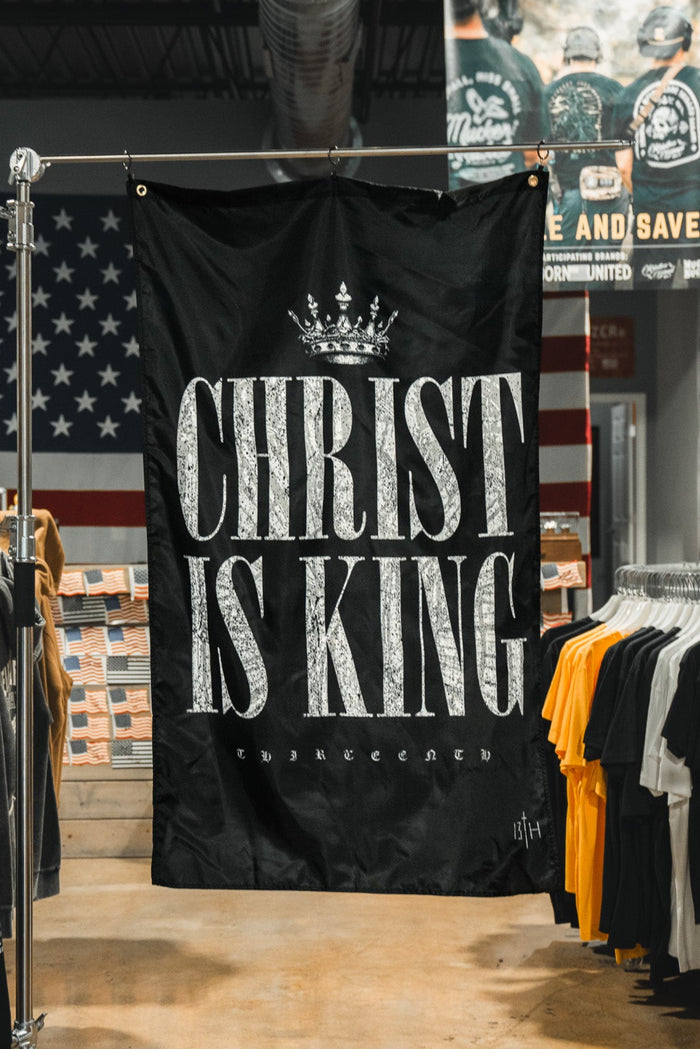 A black 13th Christ is King Flag with bold white letters hangs in a clothing store, displayed among t-shirts and a large American flag on the back wall.