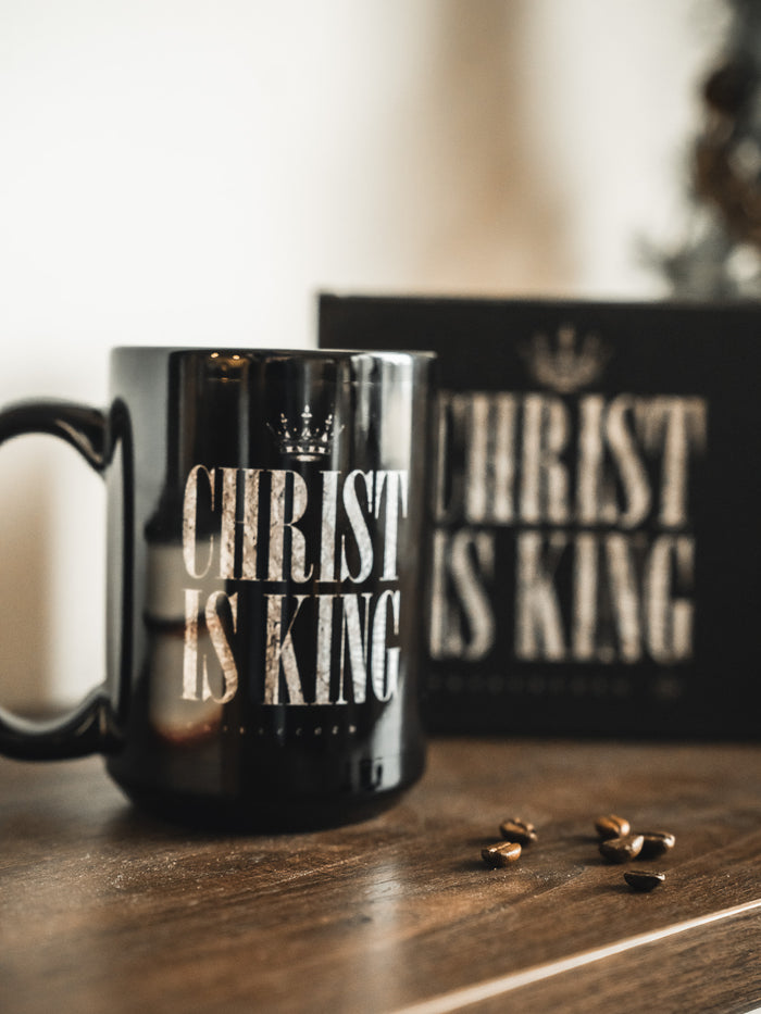 The 13th Christ is King Mug sits on a wooden surface with "CHRIST IS KING" in white letters. Coffee beans are scattered nearby, and a matching black box with the same text appears in the background.