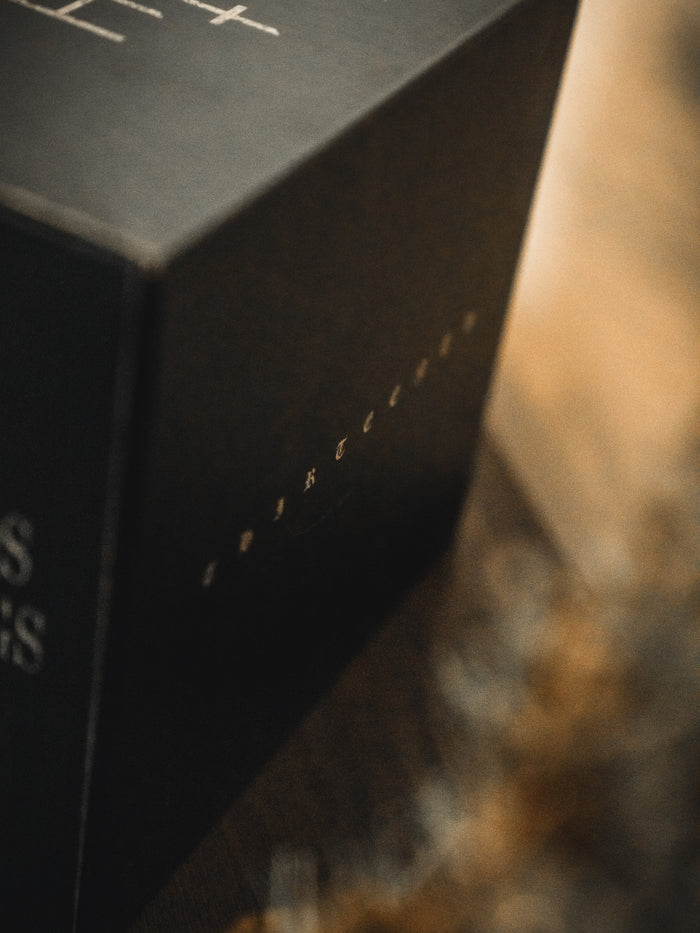 A close-up of the Christ is King Mug by 13th, showing its black edge and partially visible white "Christ is King" text, set on a wooden surface bathed in warm, soft light. Focus centers on the mug’s corner.