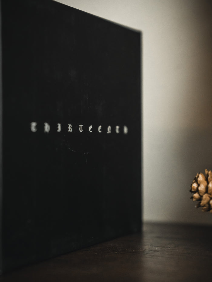A black Christ is King Mug by 13th stands upright on a dark surface. A pine cone on the right contributes to a solemn mood that echoes the mug’s bold message.