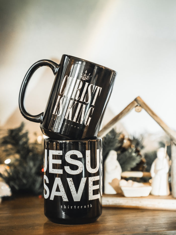 Two black 13th “Christ is King” mugs are stacked, one reading “CHRIST IS KING,” the other “JESUS SAVES.” Behind them, a nativity scene with festive greenery highlights the faith-filled message of these Christ is King Mugs.