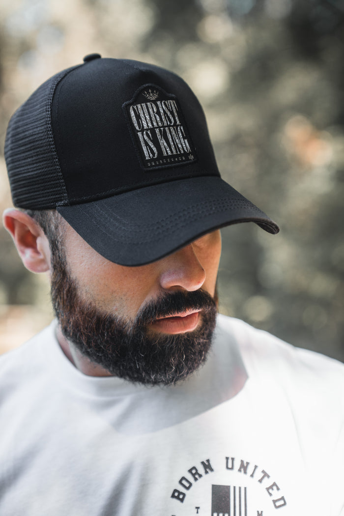 A bearded man in a white shirt with a circular "Born United" logo looks downward, wearing the 13th Christ is King Trucker Snapback. The cap is black mesh with bold lettering, and he stands against a blurred outdoor background.