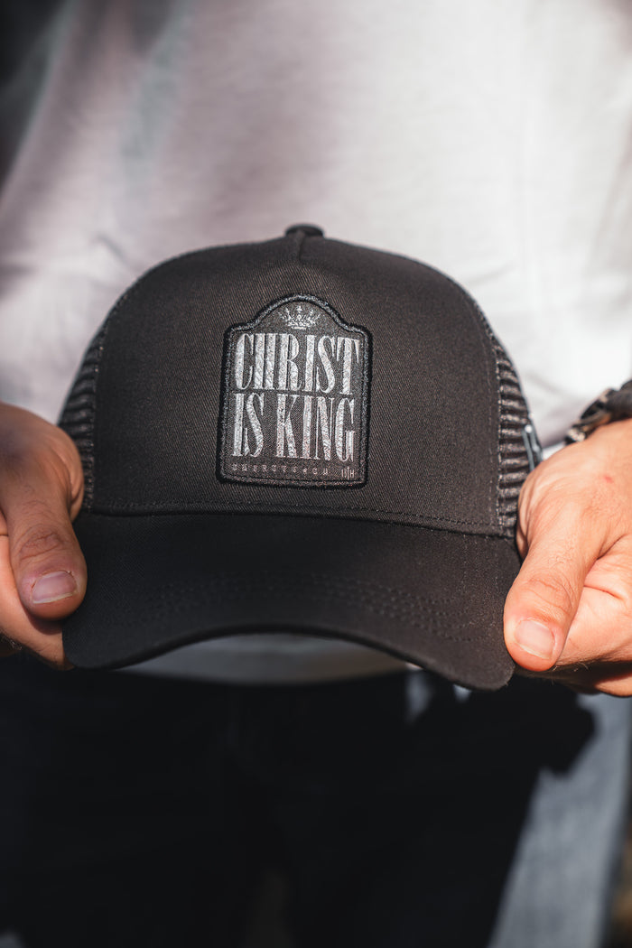 A person holds a 13th Christ is King Trucker Snapback, featuring a black handmade design with a bold white front patch. They wear a white shirt and dark pants.