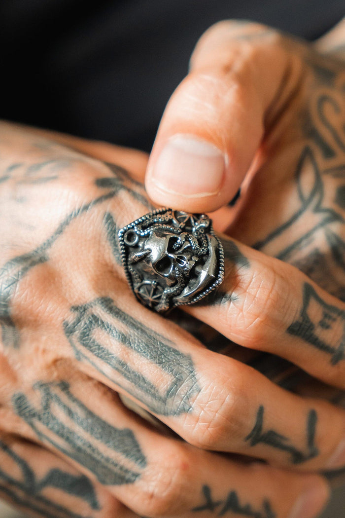 A tattooed hand sports the Relentless Betrayals Dead Men Tell No Tales Stainless Steel Ring, dark and skull-themed, exuding a first mate quality. The hand-forged piece contrasts strikingly against the skin with its ornate design and rich texture.