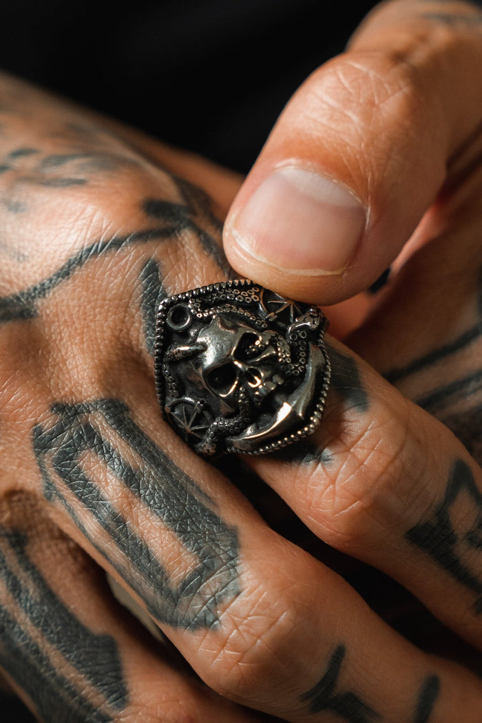 A tattooed hand showcases the Dead Men Tell No Tales stainless steel ring by Relentless Betrayal, featuring a skull and star design, with emphasis on both the ring and the intricate skin patterns.