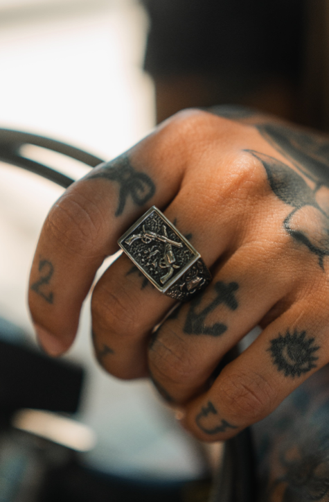 A tattooed hand, featuring an anchor and numbers, wears the Relentless Betrayal Dead or Alive Ring—a large, ornate rectangular stainless steel piece with a raised design. The softly focused, well-lit image highlights the ring’s details.