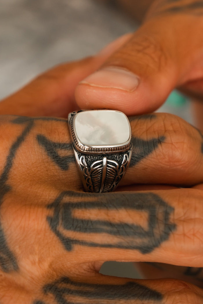 A hand with black geometric tattoos wears the Relentless Betrayal Desire Opal Ring, featuring a large, smooth white stone. The visible thumb and fingers highlight the handcrafted jewelry’s intricate details.