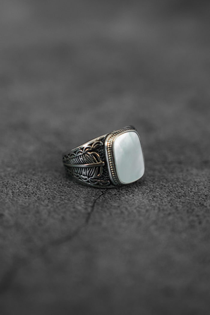 The Desire Opal Ring by Relentless Betrayal features an engraved silver band and a large, rectangular white opal, displayed on a textured dark gray surface—a stunning piece of handcrafted jewelry.