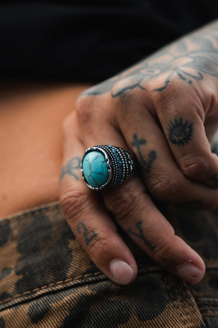 A tattooed hand rests on patterned fabric, showcasing the Relentless Betrayals Eye of the Storm Ring. This handcrafted piece features a striking turquoise stone with intricate details. The anchor and small sun tattoos enhance the allure.
