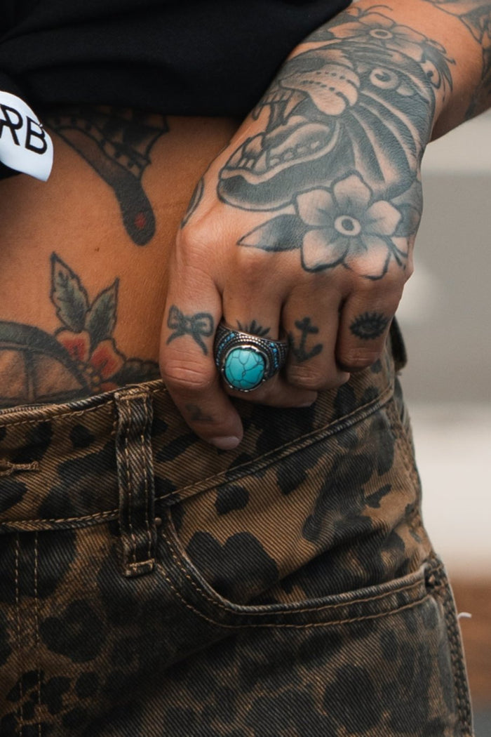 A person with floral and cross tattoos on their hand and stomach rests a hand in the pocket of leopard print pants. They wear Relentless Betrayals handcrafted Eye of the Storm Ring, its intricate design complementing a large turquoise piece on another finger.