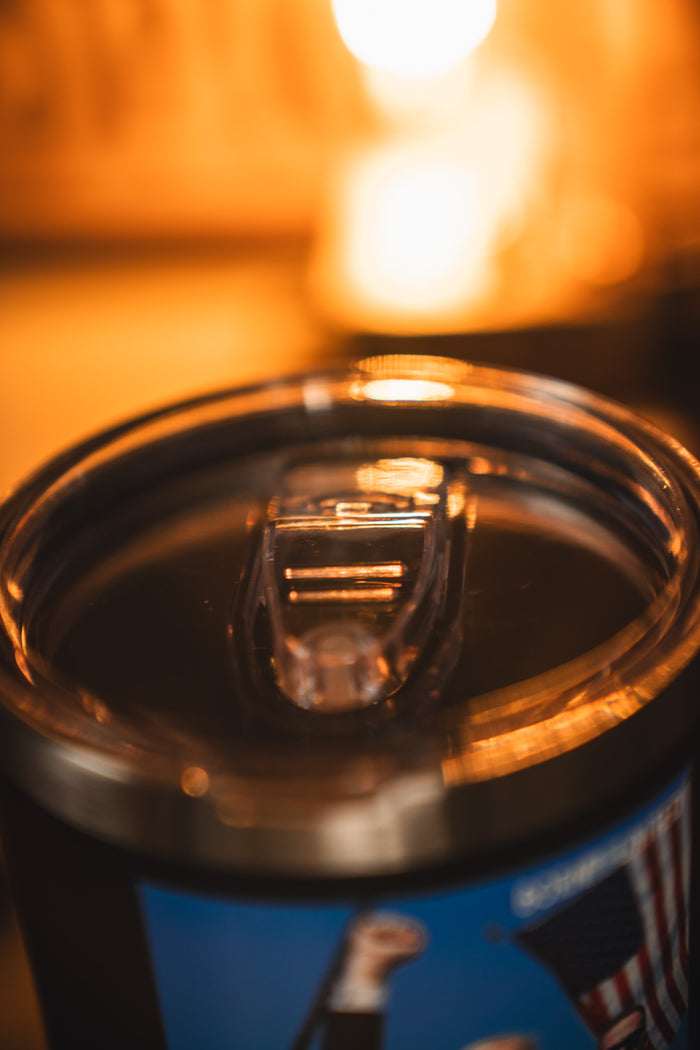 Close-up of the Born United Private Label Forever Legend Tumbler - 30oz, filled with a dark liquid and topped with a clear plastic lid. The background is blurred, glowing with warm yellow light.