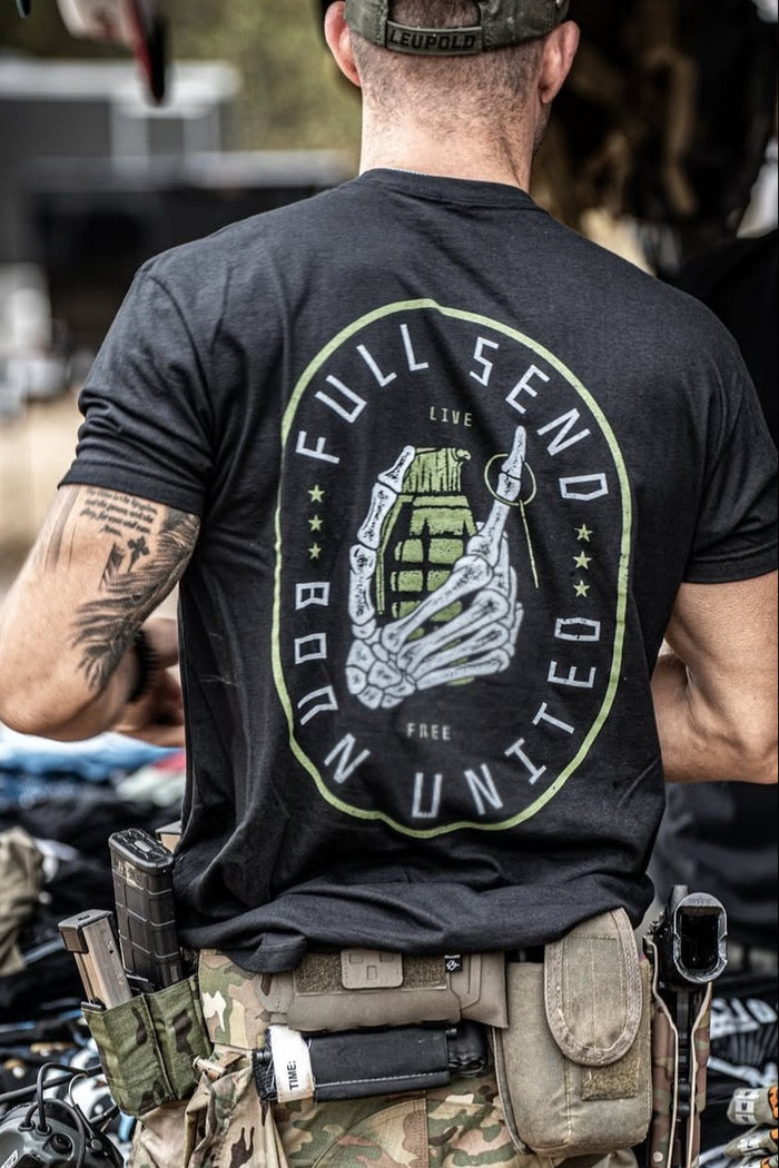 A person in a black Full Send T-shirt by Born United Private Label, featuring a bold skeleton hand design, and camouflage pants stands with their back to the camera. Their belt is loaded with tactical gear like magazine pouches and tools, enhancing the hardcore vibe of their outfit.