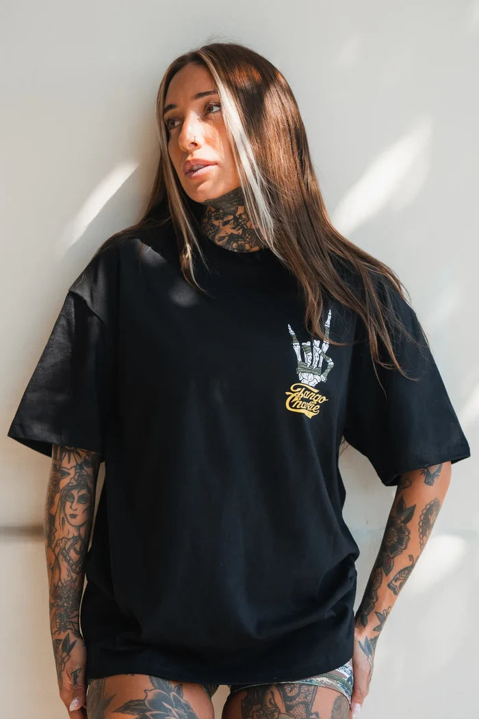 A woman with long brown hair and tattoos wears the Tango Charlie Apparel Gain Gang Premium Oversized Tee, featuring white and yellow chest graphics, as she stands against a light wall looking to the side.