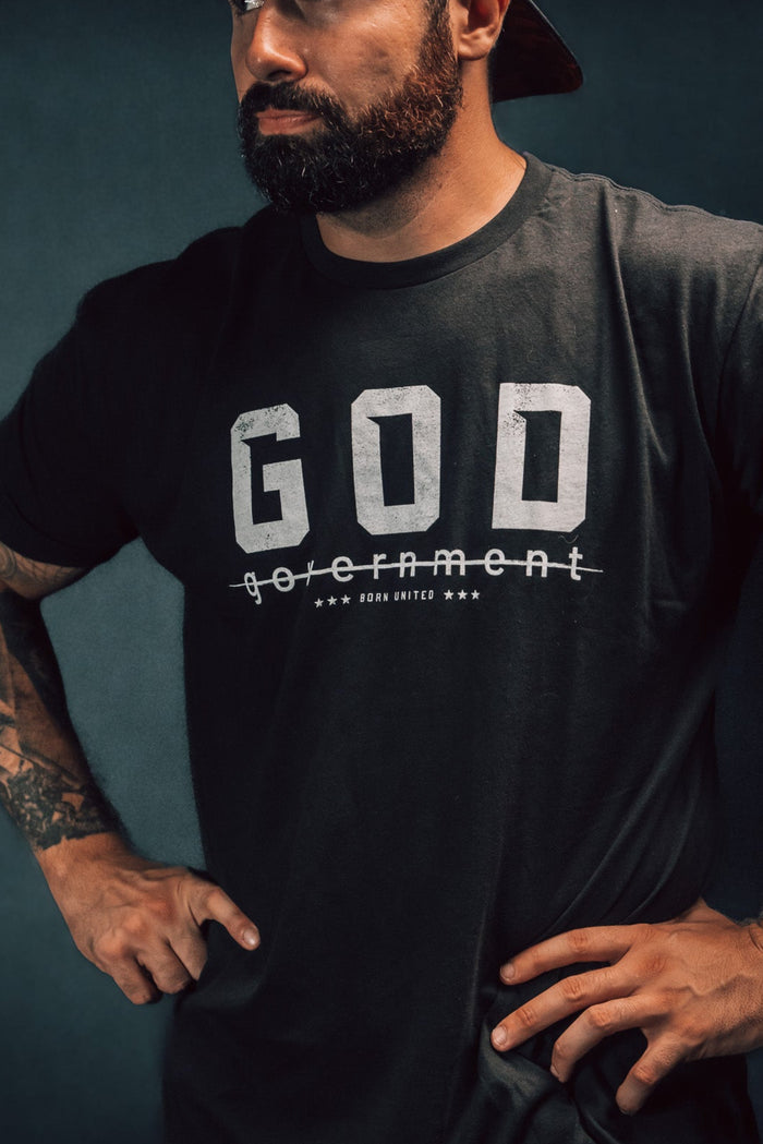 A bearded man, gazing to the side against a dark background, stands confidently with hands on hips. Hes wearing a reversed baseball cap and Born United Private Labels God Over Government t-shirt, which features the word government crossed out below GOD, challenging authority with his defiant posture.
